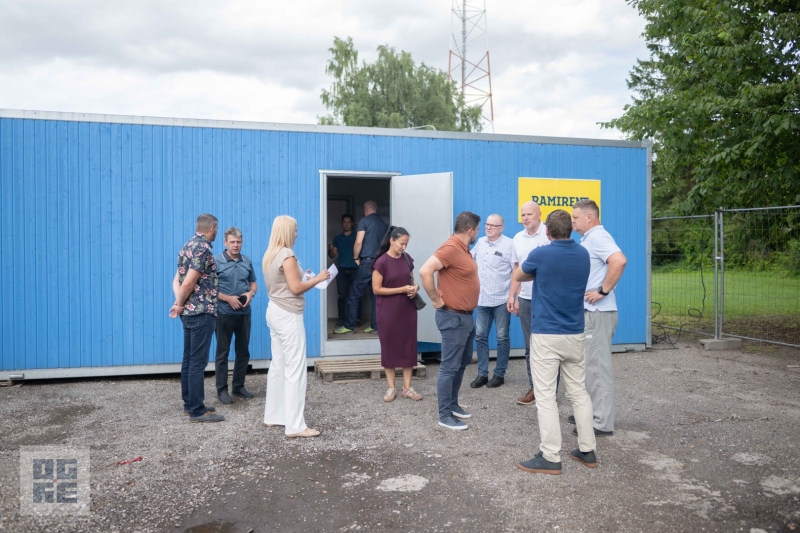 Pašvaldības un būvnieku pārstāvji, stāvot pie būvlaukuma konteinera, pārrunā autoceļa A6 pārbūves gaitu Ikšķilē 