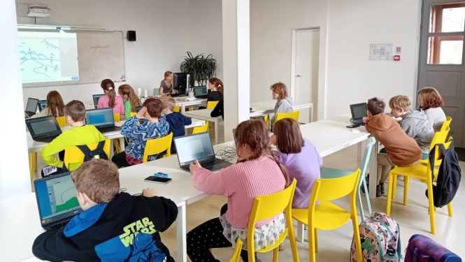 Aicinājums pieteikties Rīgas Tehniskās universitātes Bērnu un jauniešu universitātes interešu izglītības programmām Ogrē. Attēlā bērni sēž pie galdiem uz dzelteniem krēsliem. Viņiem priekšā digitālā tāfele