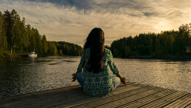 Sieviete sēž uz laipas ar skatu uz ūdeni un meditē