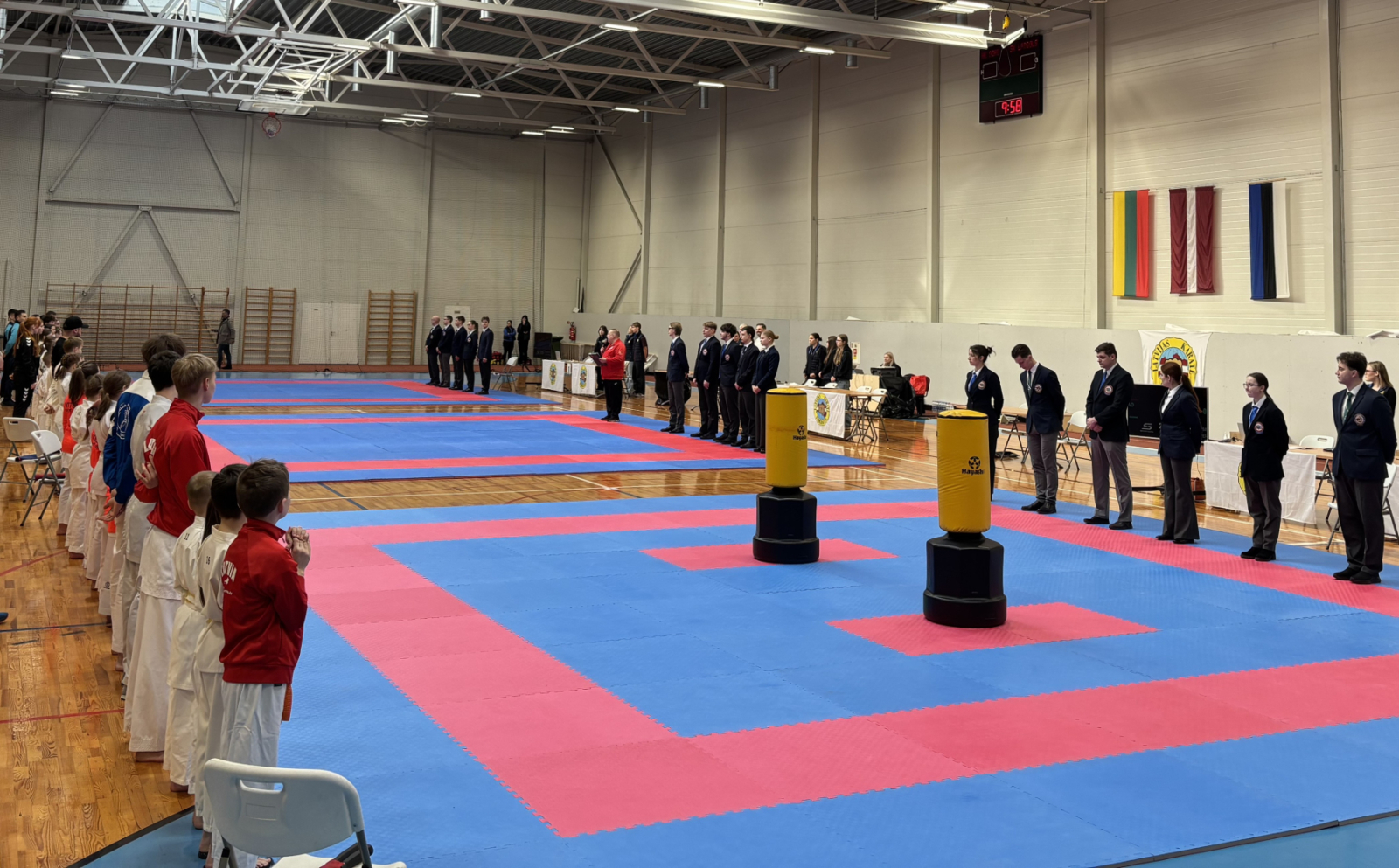 Ogrē norisinājās 24. Starptautiskais karatē turnīrs „Ogre Karate Cup & Games 2026”. Atklāšana. Laukuma vienā pusē stāv tiesneši, otrā - dalībnieki.