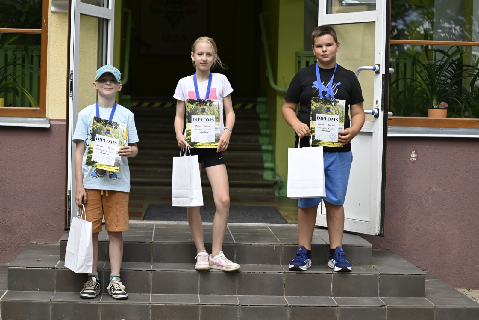 Trīs bērni ar saņemtajām godalgām uz skolas kāpnēm