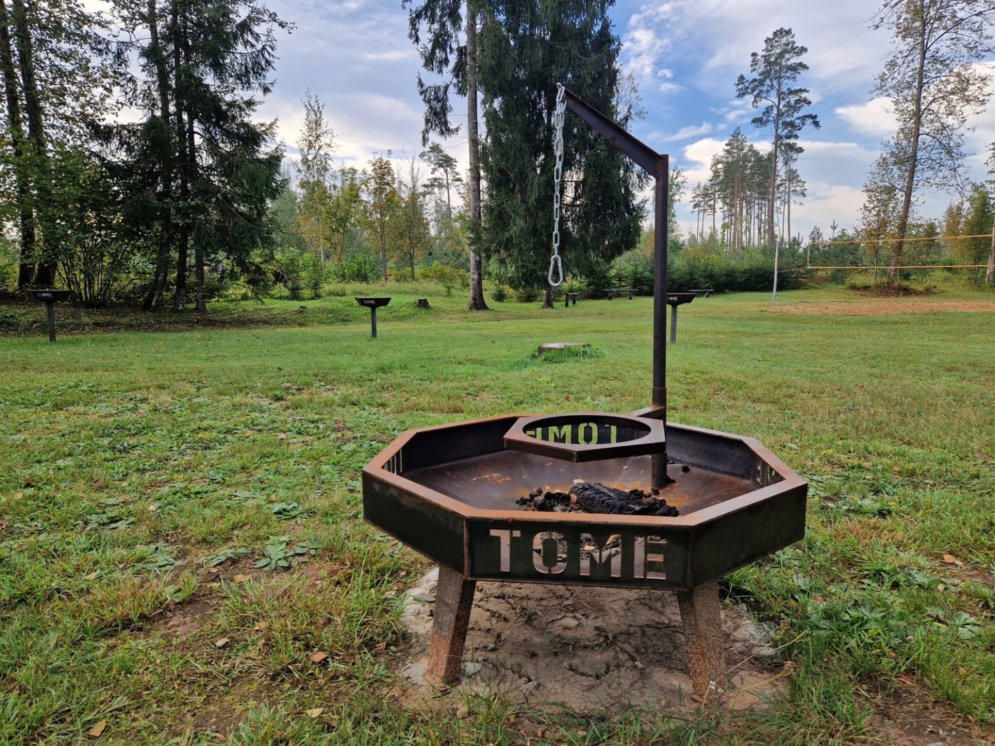 Zālē novietota metāla ugunskura vieta ar katla paliktni un uzrakstu "Tome"