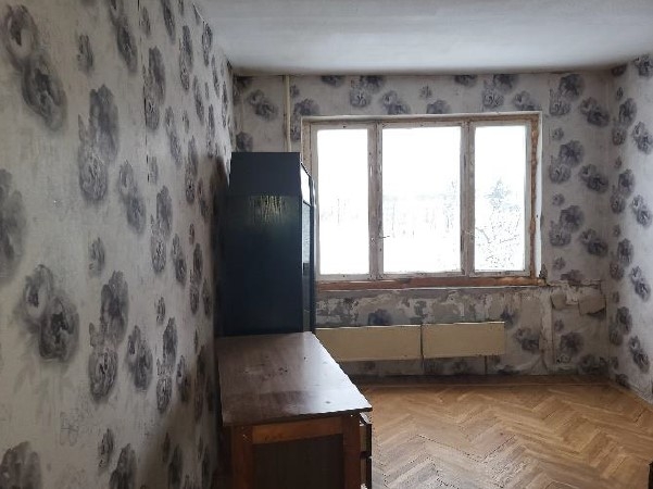 Istaba pelēkos toņos ar lielu ziedu raksta tapetēm, logu un dažām mēbelēm