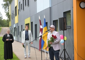 Vīrietis runā pie mikrofona pie pelēkas ēkas 23.08.2024.