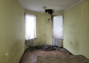 Istaba, gaiši dzeltenas, nopelējušas sienas, logs, brūna grīda