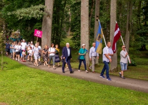 Cilvēki iet svētku gājienā, priekšā divi vīrieši ar Latvijas un Taurupes pagasta karogu Taurupes pagasta svētkos 07.2024.