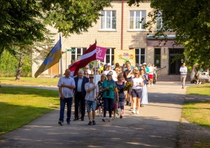Cilvēki iet svētku gājienā, priekšā valsts karogs Taurupes pagasta svētkos 07.2024.