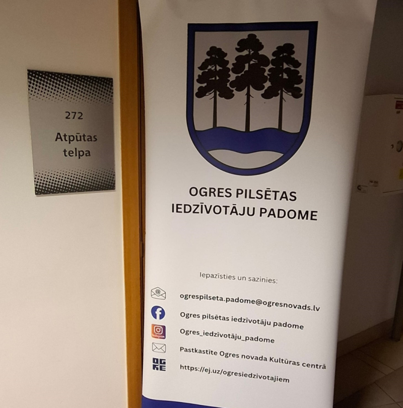 Pie 272. telpas ieejas durvīm novietots informatīvs stends ar Ogres pilsētas iedzīvotāju padomes kontaktinformāciju