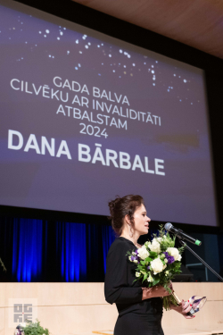 Ogres novada pašvaldības izpilddirektora vietniece Dana Bārbale ar saņemto balvu un ziediem, fonā - ekrāns ar slaidu par piešķirto balvu