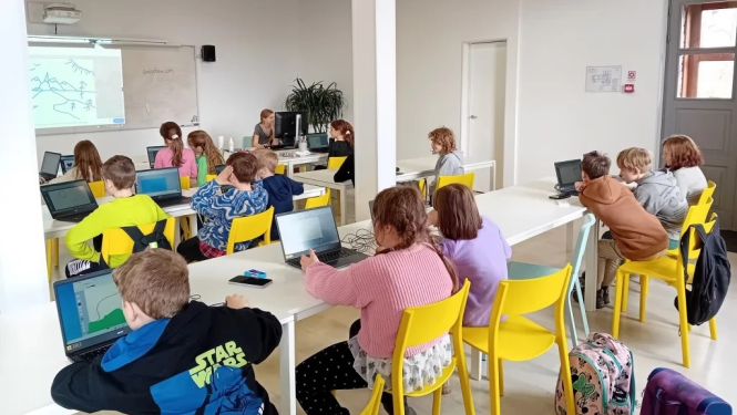 Aicinājums pieteikties Rīgas Tehniskās universitātes Bērnu un jauniešu universitātes interešu izglītības programmām Ogrē. Attēlā bērni sēž pie galdiem uz dzelteniem krēsliem. Viņiem priekšā digitālā tāfele