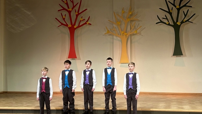 5 zēni dzied vestēs un tumšās biksēs dzied uz skatuves, fonā dekorācijās krāsaini koki 02.2024.