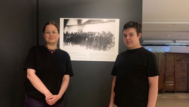 2 cilvēki melnos kreklos, gaišās biksēs stāv pie melnbaltas fotogrāfijas Ogres Vēstures un mākslas muzejā