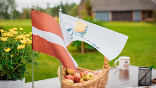 Latvijas un Vidzemes karodziņi uz lauku sētas fona