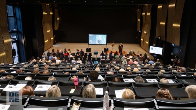 Ogres Valsts ģimnāzijas auditorijā sapulcējušies novada pedagogi uz konferenci. Fonā redzama prezentācija.