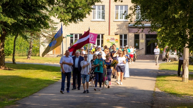 Cilvēki iet svētku gājienā, priekšā valsts karogs Taurupes pagasta svētkos 07.2024.