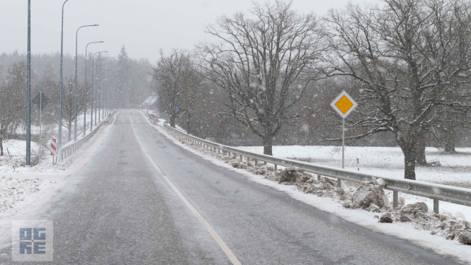 Asfaltēts ceļš Tīnūžos putenī ar norobežojošām metāla barjerām un ceļa zīmi "Galvenais ceļš"