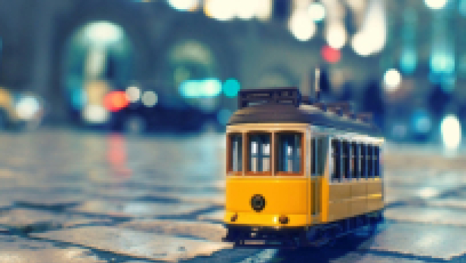 Latvijas valstspilsētās CSP aptaujās iedzīvotājus par dzīves kvalitāti. Stilizēts dzeltens tramvaja vagoniņš uz slapja ielas bruģa vakara ielu apgaismojumā