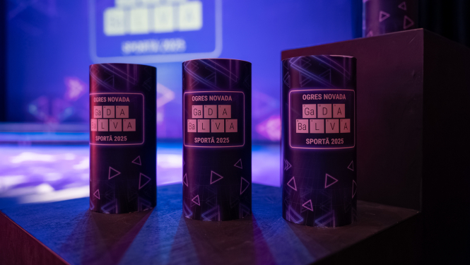 Uz podesta novietotas 3 balvas - tumši zili cilindri ar uzrakstu "OGRES NOVADA GaDA  BaLVA SPORTĀ 2025"
