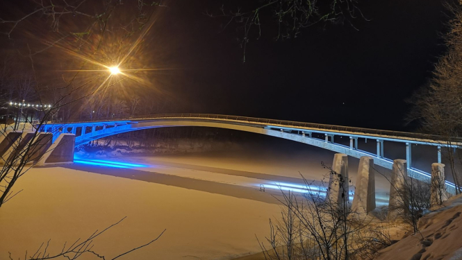 Igaunijas karoga krāsās izgaismotais arkveida gājēju tilts pār aizsalušo Ogres upi