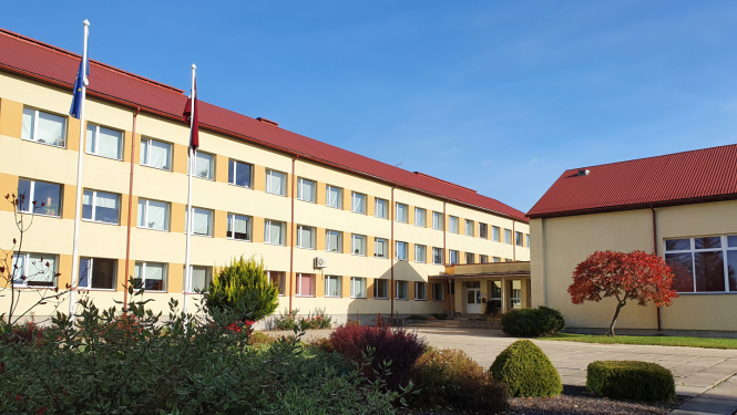 Skolas ēka ar diviem karogu mastiem un dekoratīvajiem stādījumiem priekšplānā