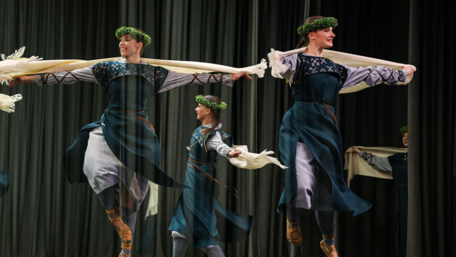 Dejojošas jaunietes zili baltos tērpos, pastalās un ar zaļiem vainagiem