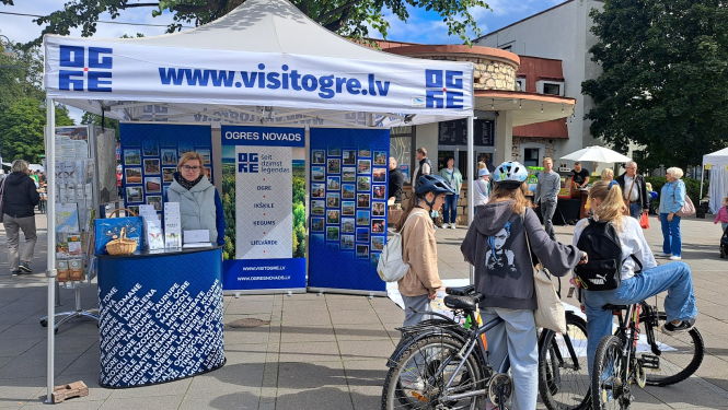Visit Ogre tūrisma informācijas stends pasākuma laikā Ogres pilsētvidē. Stendā atrodas darbinieks. Priekšplānā jaunieši uz velosipēdiem.