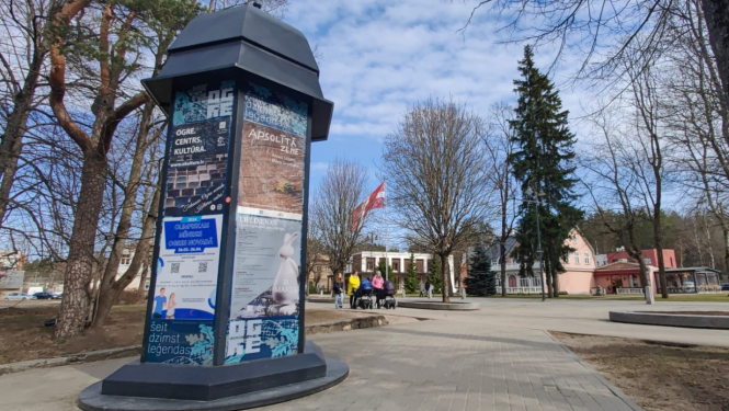 Afišu stabs Ogres centrā, fonā Latvijas karogs un māmiņas ar ratiņiem