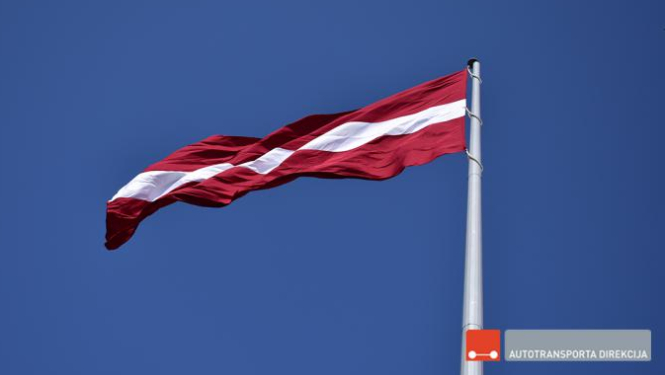 Latvijas Valsts sarkanbaltsarkanais karogs plīvo mastā uz zilu debesu fona, Autotransporta direkcijas logo uz attēla labajā apakšējā stūrī