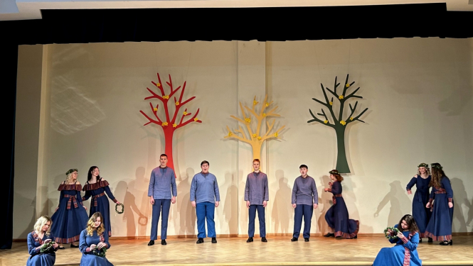 Jaunieši zilos tērpos dzied uz skatuves, fonā dekorācijās krāsaini koki 02.2024.