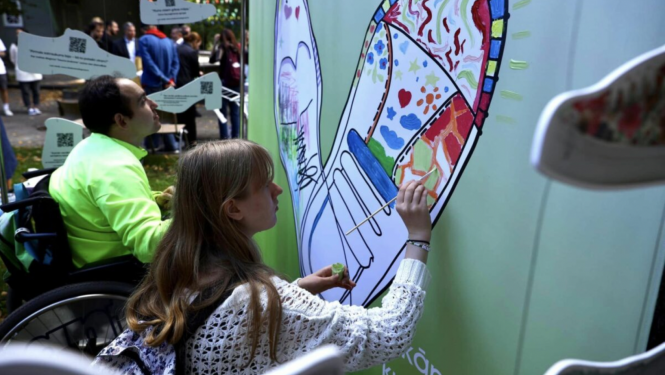 Vīrietis un sieviete ratiņkrēslos zīmē krāsainas kurpes uz liela zaļa audekla