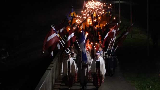 Cilvēki, priekšplānā tautas tērpos, dodas gājienā ar Latvijas karogiem un lāpām pa Ogres molu par godu Lāčplēša dienai
