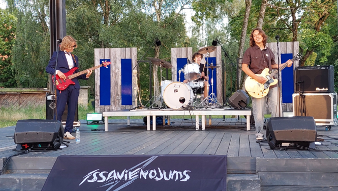 Grupas "Īssavienojums" trīs dalībnieki uzstājas uz Ogres pilsētas skvēra skatuves, priekšplānā uzraksts "Īssavienojums"