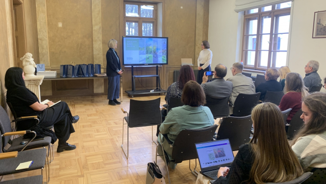 Pasākuma norises zāles kopskats ar dalībnieku sēdvietu rindām, ekrānu un divām studentēm, kas sniedz prezentāciju