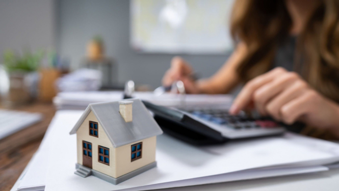 Kartona māja, fonā sieviete ar kalkulatoru