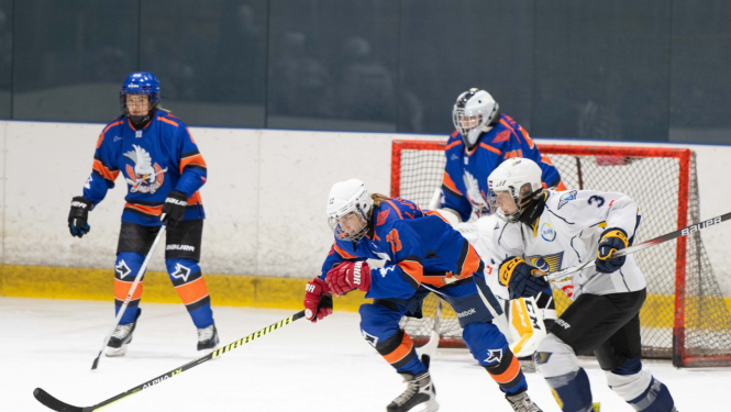 Trīs sieviešu hokeja komandas "Ogres Grācijas" spēlētājas zilos formas tērpos spēles laikā uz ledus sacenšas par ripu ar pretinieču komandas spēlētājām