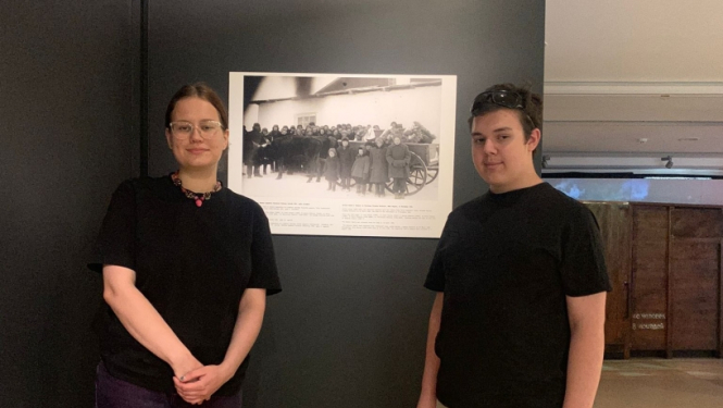 2 cilvēki melnos kreklos, gaišās biksēs stāv pie melnbaltas fotogrāfijas Ogres Vēstures un mākslas muzejā