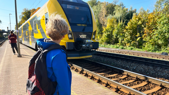 Jaunietis zilā jakā ar mugursomu stāv pie vilciena