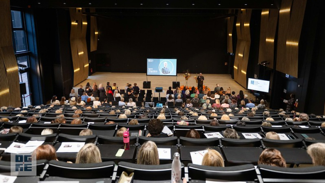 Ogres Valsts ģimnāzijas auditorijā sapulcējušies novada pedagogi uz konferenci. Fonā redzama prezentācija.