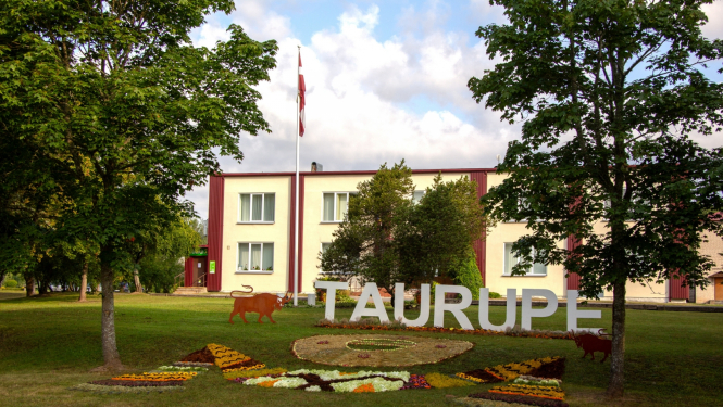 Uzraksts Taurupe ar ziediem, fona daudzstāvu māja Taurupes pagasta svētkos 07.2024.