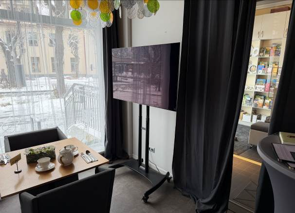 Kafejnīcas zāle un uz statīva novietots ekrāns pie zāles izejas