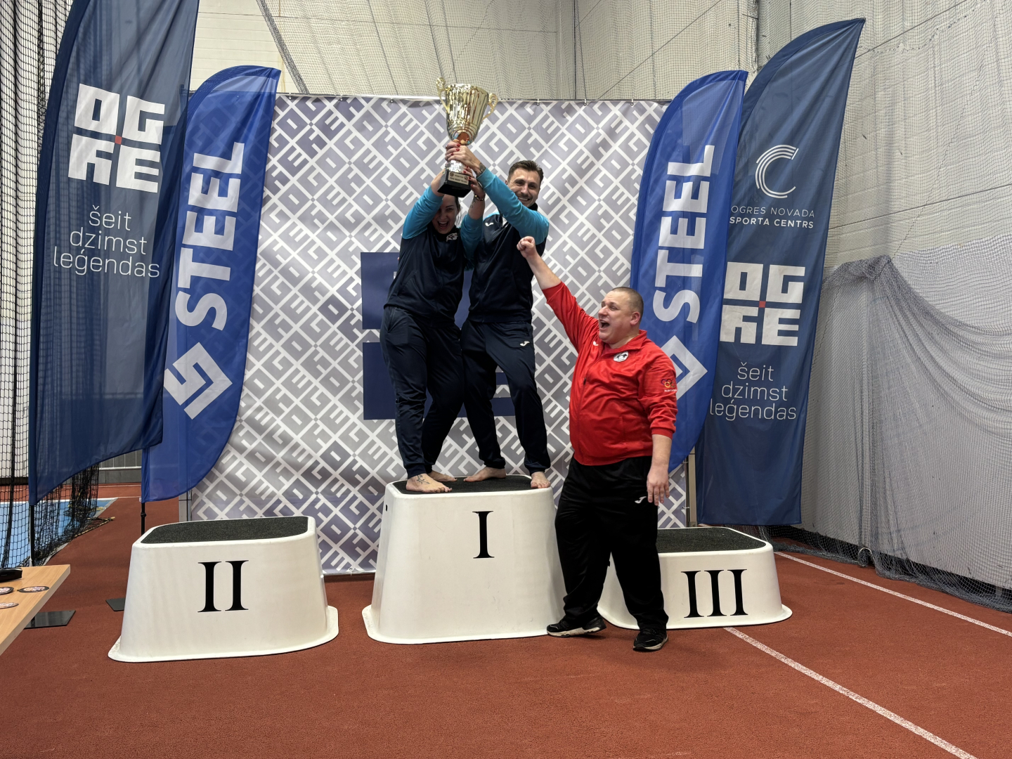 24. Starptautiskais karatē turnīrs „Ogre Karate Cup & Games 2026”. Uz pjedestāla pirmās vietas divi uzvarētāji, kuriem rīkotājs pasniedz kausu. 