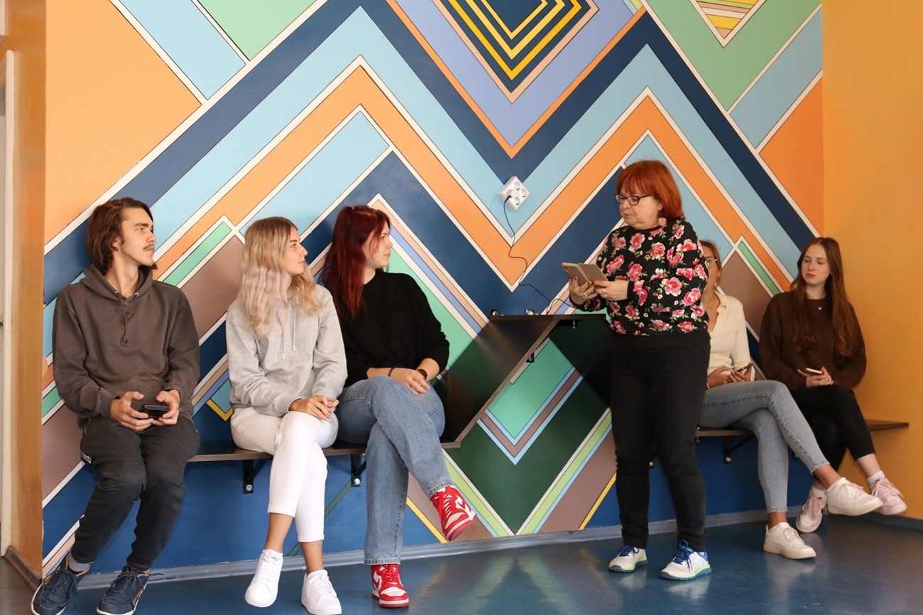 5 skolēni sēž uz koka soliem, kas uzstādīti pie sienas gleznojuma, un klausās skolotājā