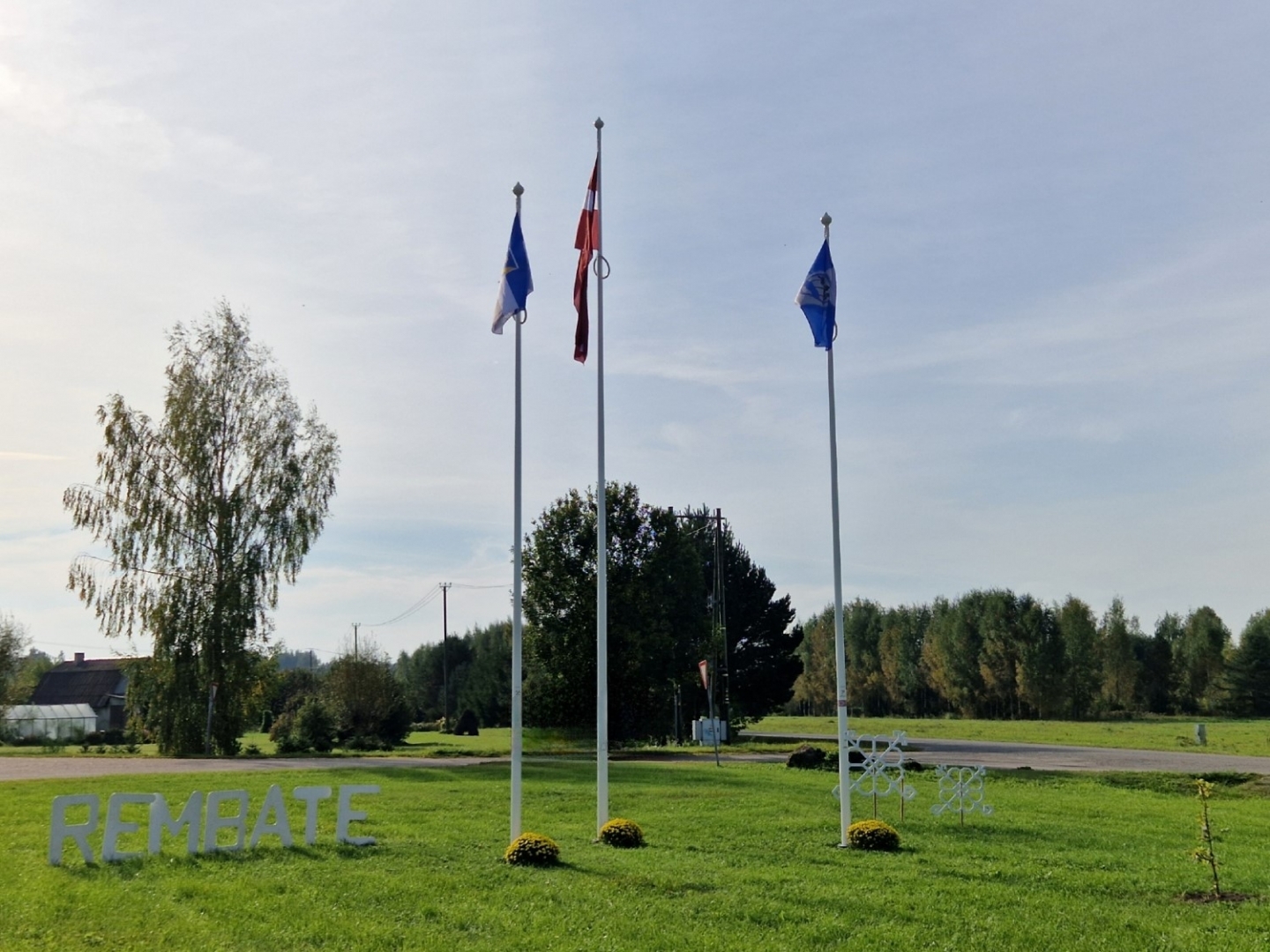 3 karogu masti ar Latvijas, Ogres novada un Rembates karogiem un zālē izvietotu uzrakstu Rembate