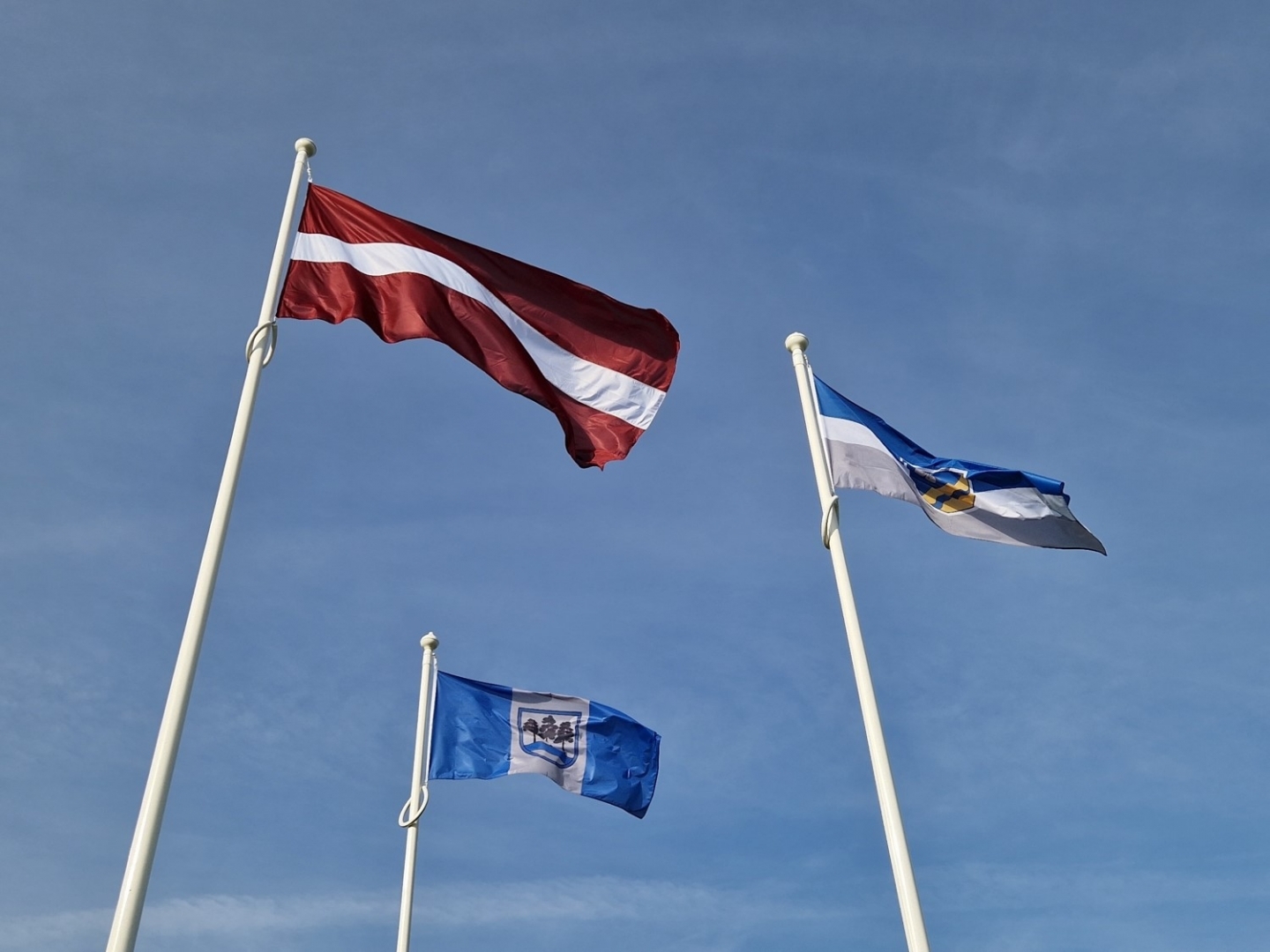 Uz debesu fona vējā plīvo Latvijas, Ogres novada un Rembates pagasta karogi