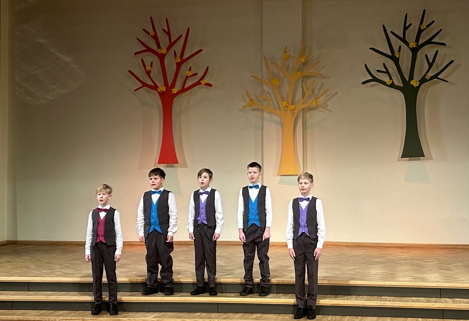 5 zēni dzied vestēs un tumšās biksēs dzied uz skatuves, fonā dekorācijās krāsaini koki 02.2024.