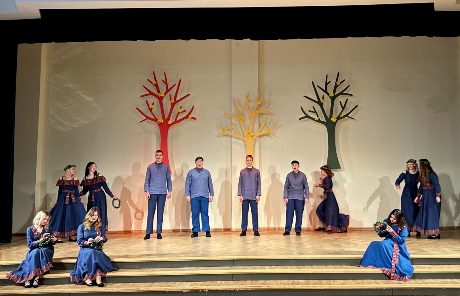 Jaunieši zilos tērpos dzied uz skatuves, fonā dekorācijās krāsaini koki 02.2024.