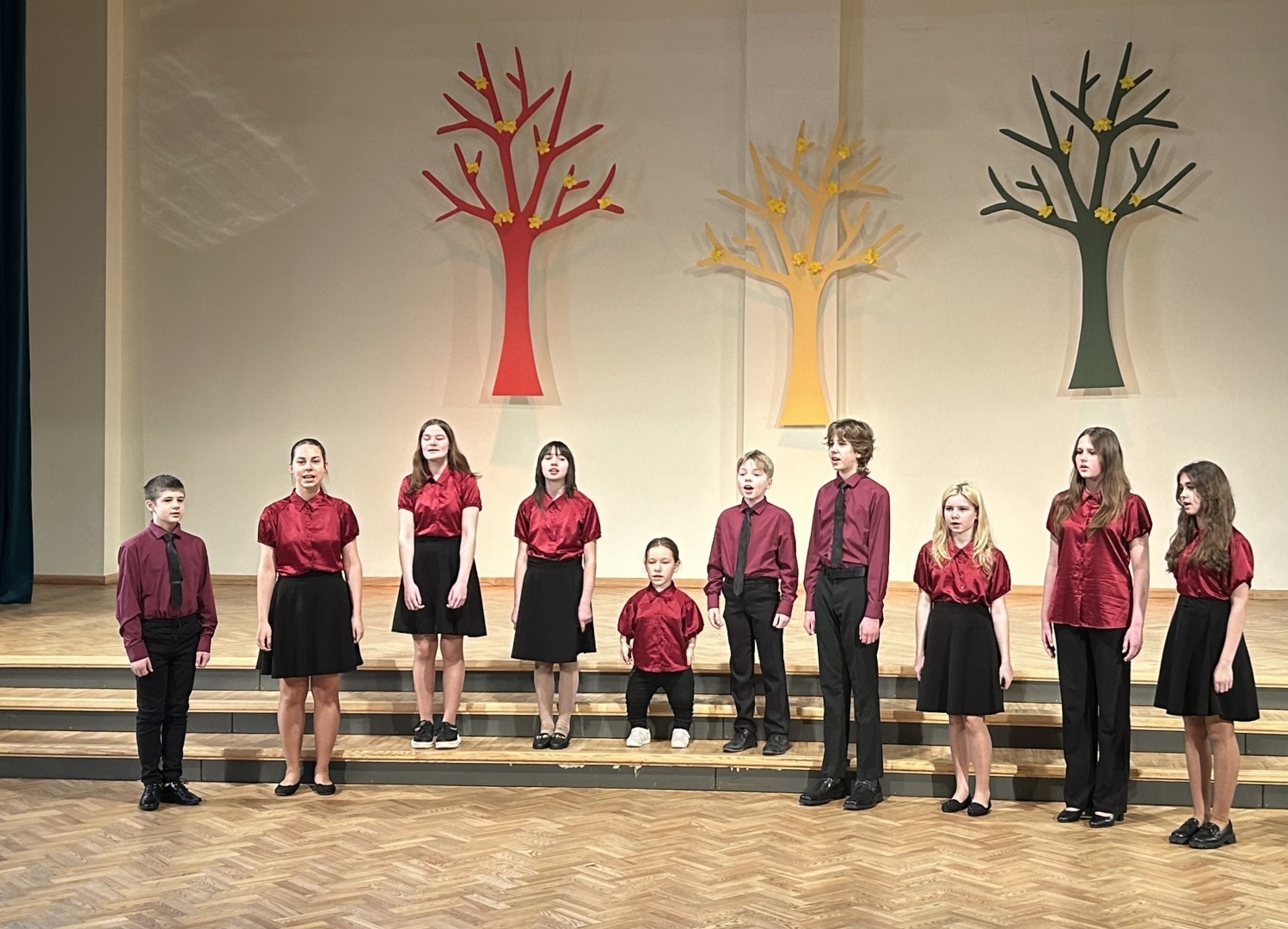 10 bērni sarkanās blūzēs un kreklos, tumšās biksēs un svārkos dzied uz skatuves, fonā dekorācijās krāsaini koki 02.2024.