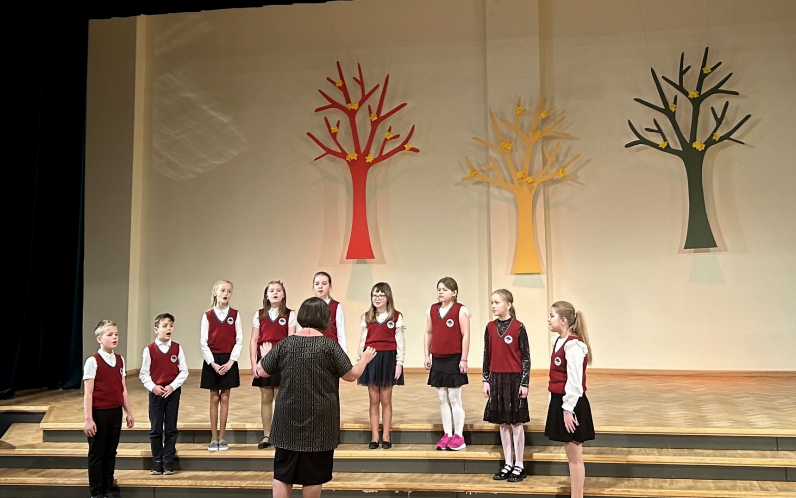 9 bērn Ogresgala pamatskolas formas tērpos - sarkanas vestes, baltas blūzes, melni svārki dzied uz skatuves, diriģē skolotāja. Fonā dekorācijās krāsaini koki 02.2024.
