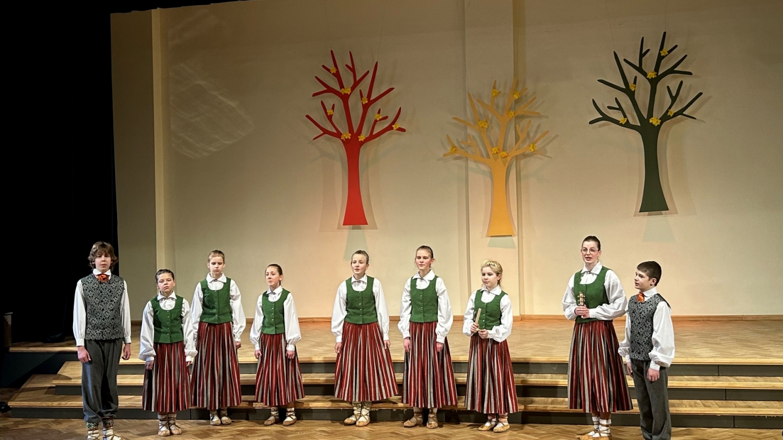 Bērni tautas tērpos dzied uz skatuves, fonā dekorācijās krāsaini koki 02.2024.