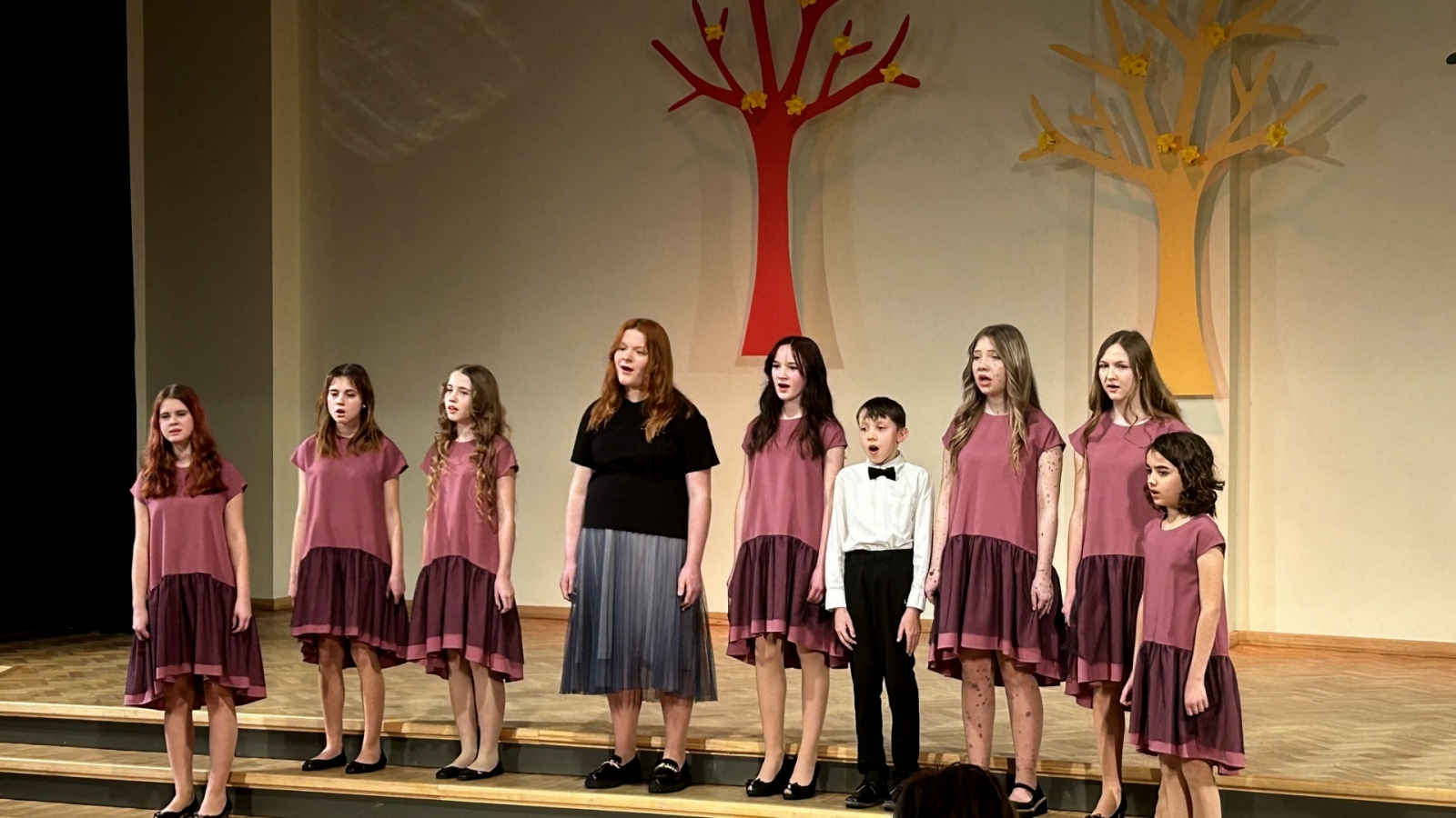 t meitenes sarkanīgās kleitās, viena melnā kleita un viens zēns dzied uz skatuves dzied, fonā dekorācijās krāsaini koki 02.2024.